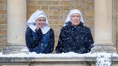 Two women wearing nun's habits stood outside in the snow next to an old, grand building. One is smiling and waving at someone out of shot