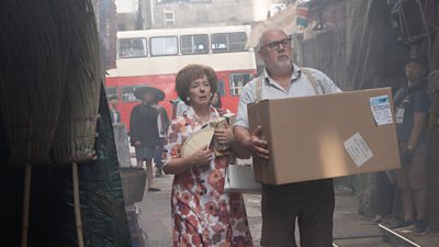 A woman in a floral dress carrying a fan and a Christmas present, walking down a street with a man who is holding a large cardboard box