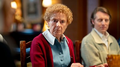 Linda Bassett in character as Nurse Phyllis wearing a red cardigan over a nurse's dress, sat at a table on a wooden chair