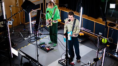 Two people are playing guitars in a recording studio, surrounded by lighting and the rig camera setup.