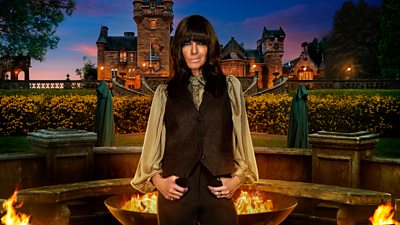 Claudia Winkleman stands by a firepit outside a castle at dusk, wearing a waistcoat and trousers and a long bell sleeve shirt.