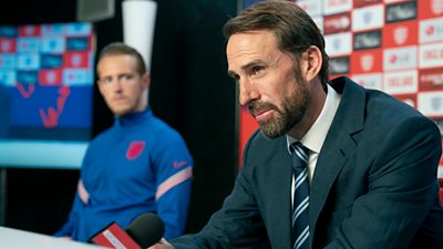 Photo of Gareth Southgate (Joseph Fiennes) and Harry Kane (Will Antenbring) in a scene from Dear England. Gareth Southgate sits in front of a microphone beside Harry Kane.
