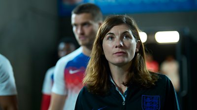 Pippa Grange (Jodie Whittaker) dressed in a fleece with the England football team branding. She walks onto a football pitch and looks up at the view. Football players in their team kit are visible in the background. 