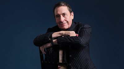 Photo of Jools Holland sat backwards on a chair in front of a deep blue background. 