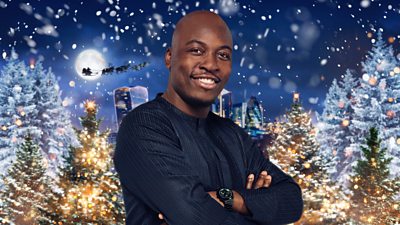 A smiling man in navy blue long-sleeved top stands outside a festive backdrop of Christmas trees 