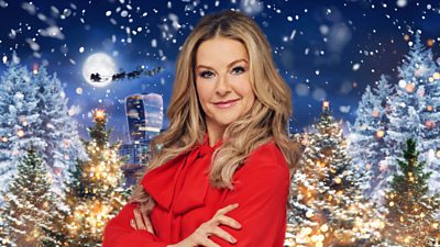 A blonde woman in a red shirt with a pussy bow poses with her arms folded, amid a festive setting of Christmas trees and snow.