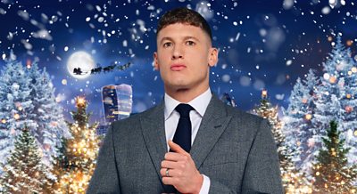 A man in a grey suit with a stylish haircut stands amid a festive setting: Christmas trees, snow and even Santa riding by on his sleigh.