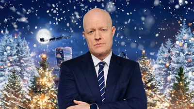 A bald man in a blue suit with his arms folded looks on as a scene of festivity takes place behind him, such as snow and Christmas trees.