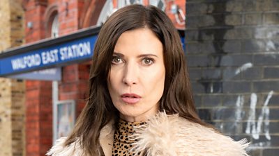 A woman in a leopard print top and fluffy jacket is standing outside the fictional Walford East tube station
