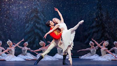 Image from a performance of The Nutcracker from the Royal Ballet. A man in a red military jacket holds a woman in a white dress. Ballerina's hold a pose in the background.
