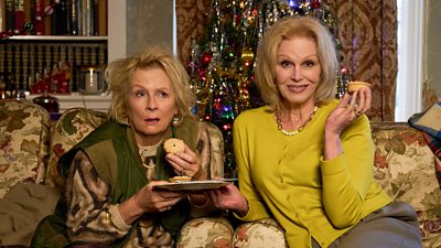Two women pose with mince pies, sitting on floral furniture in a house decorated for Christmas