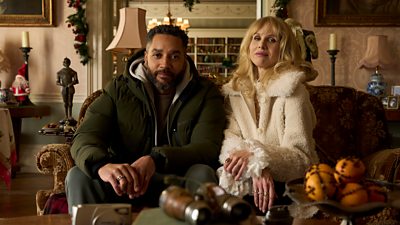 A man and woman sit side by side on a sofa in winter coats