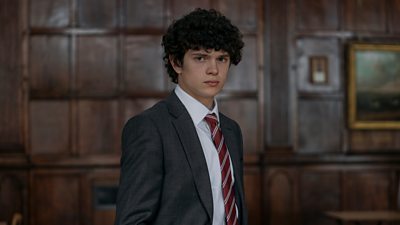 A boy in school uniform looks to camera, standing in a wood panelled room