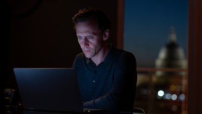 A man sits at a laptop at night, typing in the dark, his face reflected in the glow of the screen. The dome of a large building is visible through a window behind him.
