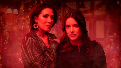 Key art for the Christmas Day episode of EastEnders. Characters Kat and Zoe stand in a festively decorated pub. A red filter has been applied to the image making the scene appear ominous.