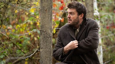 A man in a brown coat stands in the woods looking worried, reaching into his coat for something