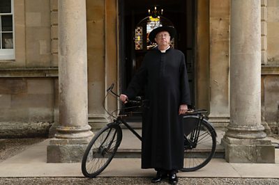 A priest (played by Mark Williams) looks on solemnly with a bicycle.