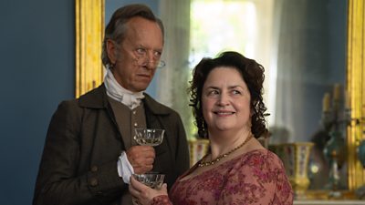 A man and a woman hold wine glasses and stand beside a large mirror in a grand house. They both wear Regency-era clothing. 