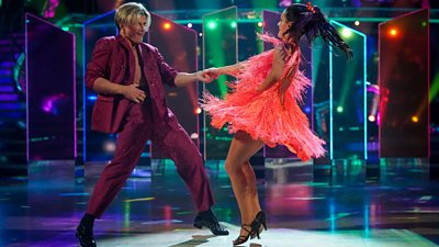 A man and woman dance a Jive at speed. He wears a purple suit and she wears a bright pink fringe dress.