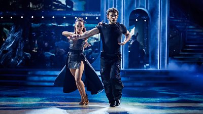 A man and a woman dance a Paso Doble. They wear dark clothing as they march across a dark studio floor.