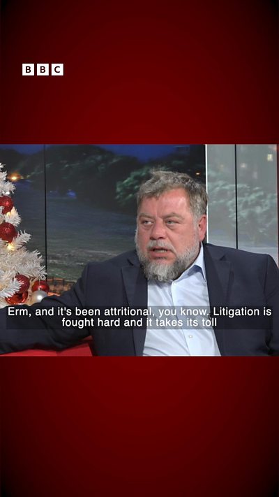 A bearded man, wearing a dark suit and white shirt, sits in the BBC Jersey television studio