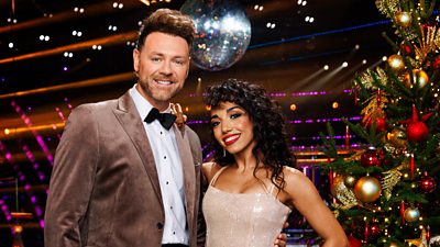 Brian McFadden and Michelle Tsiakkas stand together smiling in the Strictly ballroom, standing beside a decorated Christmas tree with a glittering disco ball visible behind them