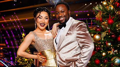 Babatunde Aléshé and Nancy Xu stand together smiling in the Strictly ballroom, standing beside a decorated Christmas tree with a glittering disco ball visible behind them
