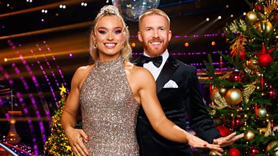 Jodie Ounsley and Neil Jones stand together smiling in the Strictly ballroom, standing beside a decorated Christmas tree with a glittering disco ball visible behind them