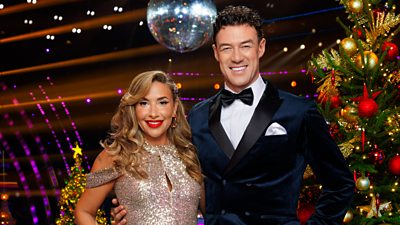 Melanie Blatt and Kai Widdrington stand together smiling in the Strictly ballroom, standing beside a decorated Christmas tree with a glittering disco ball visible behind them