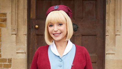 Helen George in character as Trixie Aylward. Wearing a midwife uniform, she stands on the steps to Nonnatus House.