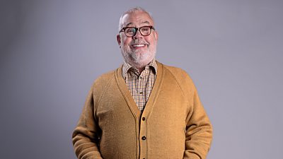 Cliff Parisi in character as Fred Buckle. He smiles to camera and wears a warm, beige cardigan over a plaid shirt. 