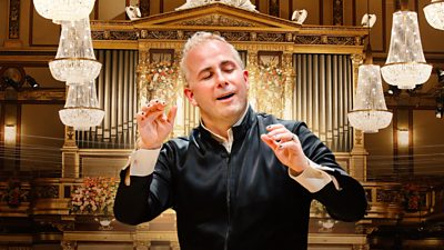 A person conducts an orchesta in an elaborate hall, where chandeliers hang from the ceiling