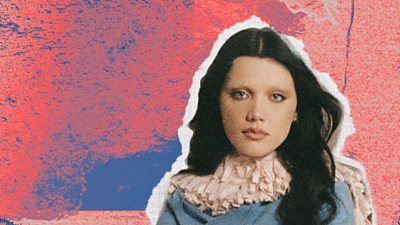 A picture of Alessi Rose on a pink and blue background. Alessi has wavy dark brown hair, and is wearing a blue top with a white ruffled collar.