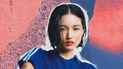 A picture of Chloe Qisha on a pink and blue background. Chloe has short cropped black hair and red lipstick, and is wearing a mid-blue polo top with white sporty stripes along the shoulders.