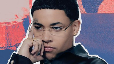 A picture of kwn on a pink and blue background. Kwn is wearing clear glasses, has a small tattoo on her forehead, and is wearing a black shirt with a collar. Her pointer finger is pressed against her temple, the rest of her fingers leaning against her face.