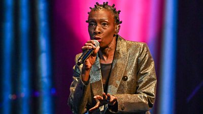 A woman (Alison Limerick) in a shimmering gold suit jacket sings on stage with a microphone in hand
