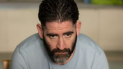 A head a shoulders shot of a man in a grey top, staring ahead looking serious
