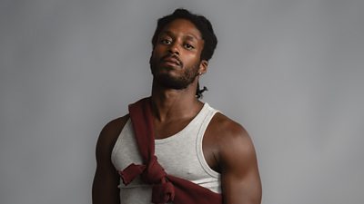A man in a grey tank top stares to camera with a red jumper tied around his shoulder