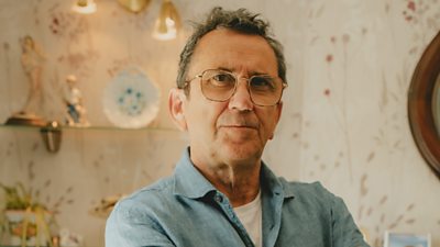 A man in a blue shirt and white t-shirt looks to camera, standing in a living room