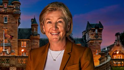 A woman in a white top and burnt organge/brown blazer smiles to camera, set against the backdrop of a castle by night