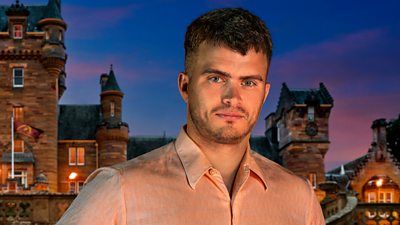 A man in a peach shirt smiles to camera, set against the backdrop of a castle by night