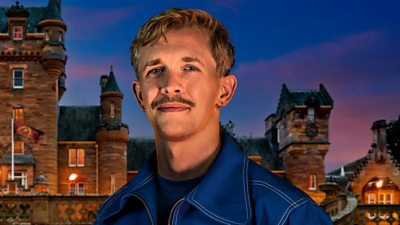 A young man with a moustache smiles to camera, wearing a dark blue jacket with white trim and a dark blue top. He's set against the backdrop of a castle by night.