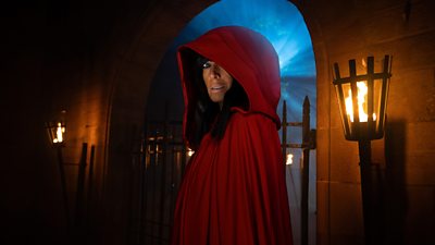 A figure in a red cloak (Claudia Winkleman) looks to camera, standing in an archway of a castle by night