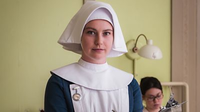 A woman wearing a nun’s midwifery uniform stands on a hospital ward. A patient sits in a hospital bed in the background. 