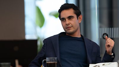 A man wearing a navy blazer over a jumper, sat in an office. Plants are visible in the background, and a glass mug of coffee sits on the desk in front of him. 