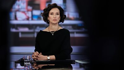 Indira Varma looks serious in a news studio with hands clasped in front of her. She is wearing a black dress, with gold necklace and watch.