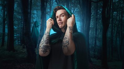 A man in a green cloak holds the hood around his face, set against a backdrop of a dark forest