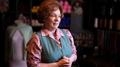 A woman dressed in a pink, floral blouse and a grey, checkered waistcoat stands indoors. Balls of cotton and fabrics are visible in the background. 