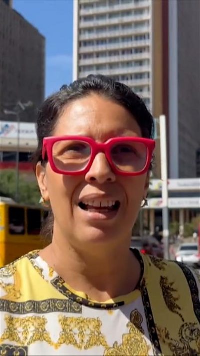 Woman wearing red glasses and a yellow printed shirt speaks towards a camera with buildings behind her.