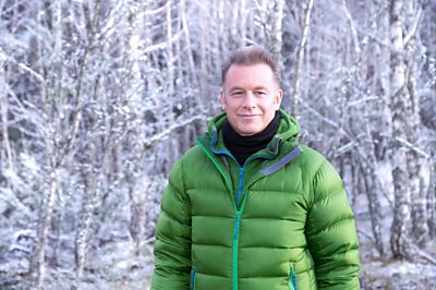 A man in a green puffy jacket (Chris Packham) stands in a wintery forest 